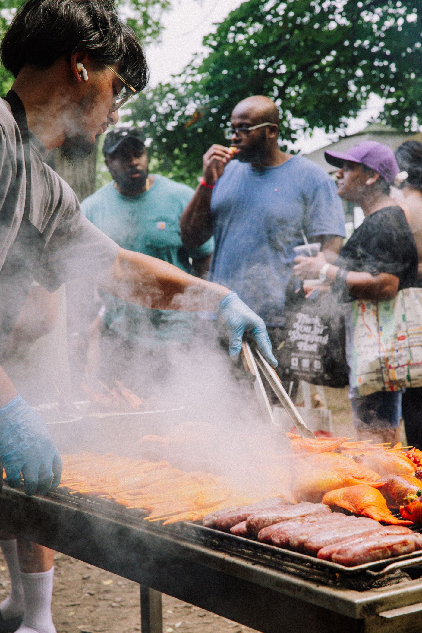 Food cooks over an outdoor barbecue grill as people stand nearby.